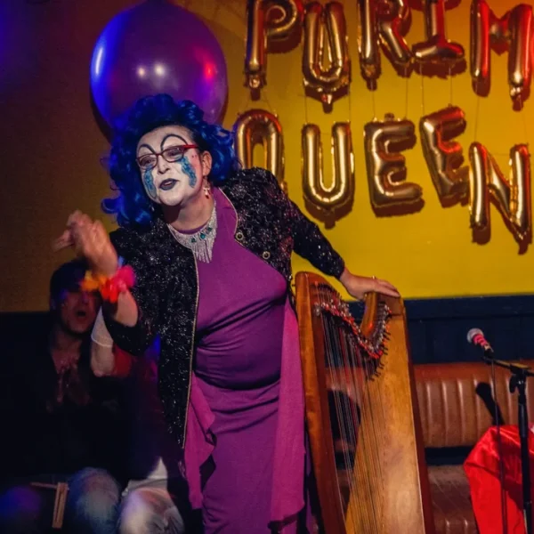 Our very own Purim Queen, Mx Bag performing the Book of Esther in a blue wig, purple gown, and clown makeup.