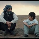 Man en kind staren elkaar zitten naast elkaar aan in de woestijn in film Neighbours van Mano Khalil