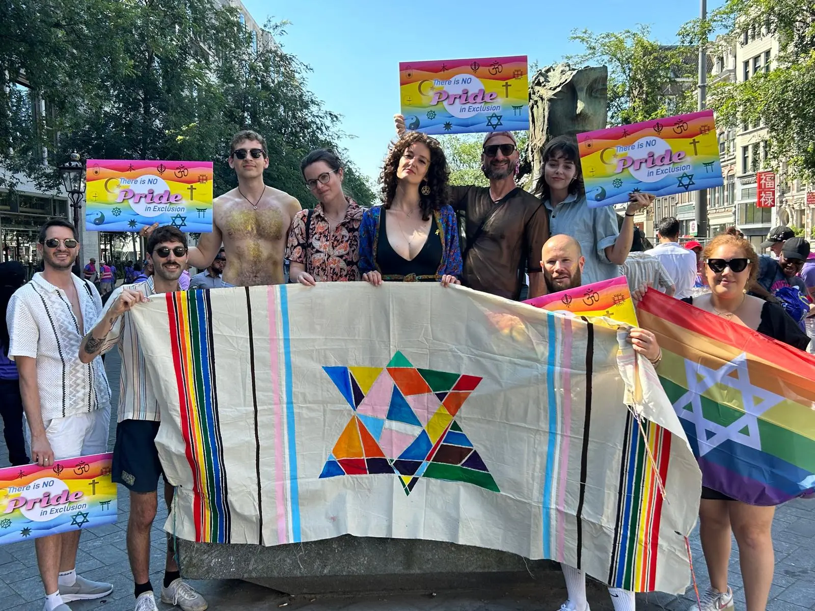 A group holds a banner for Pride March. The banner features a rainbow-colored star of David, rainbow colored tallit (prayer shawl) stripes. Their signs read "no pride in exclusion"